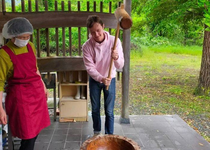 A person in a pink hoody in the process of swinging a hammer down on Japanese glutinous rice, the traditional way to make mochi.